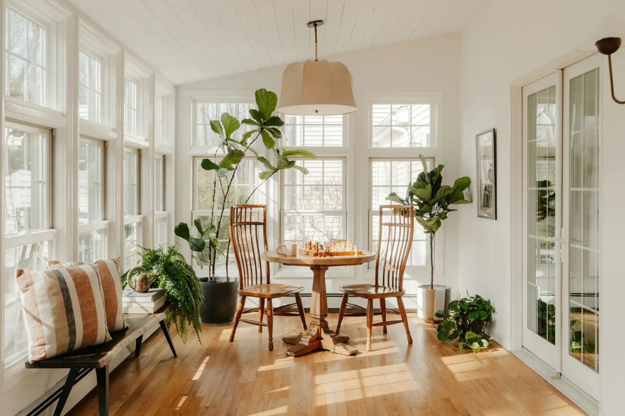 A sunlit sunroom with a round wooden table, a chess set, and potted fiddle-leaf figs